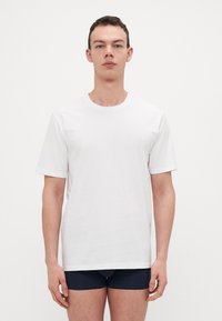 Hvit kortermet t-skjorte laget av myk bomull, med klassisk rund hals og avslappet passform. Båret med mørkeblå shorts, sett forfra.