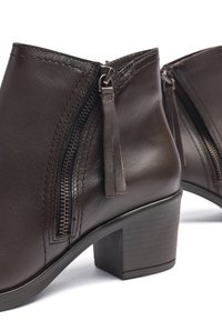 Botas de tobillo de cuero marrón con un acabado texturizado, que cuentan con cremalleras laterales, tiradores con borlas y un tacón grueso. Contornos suaves y costuras sutiles.