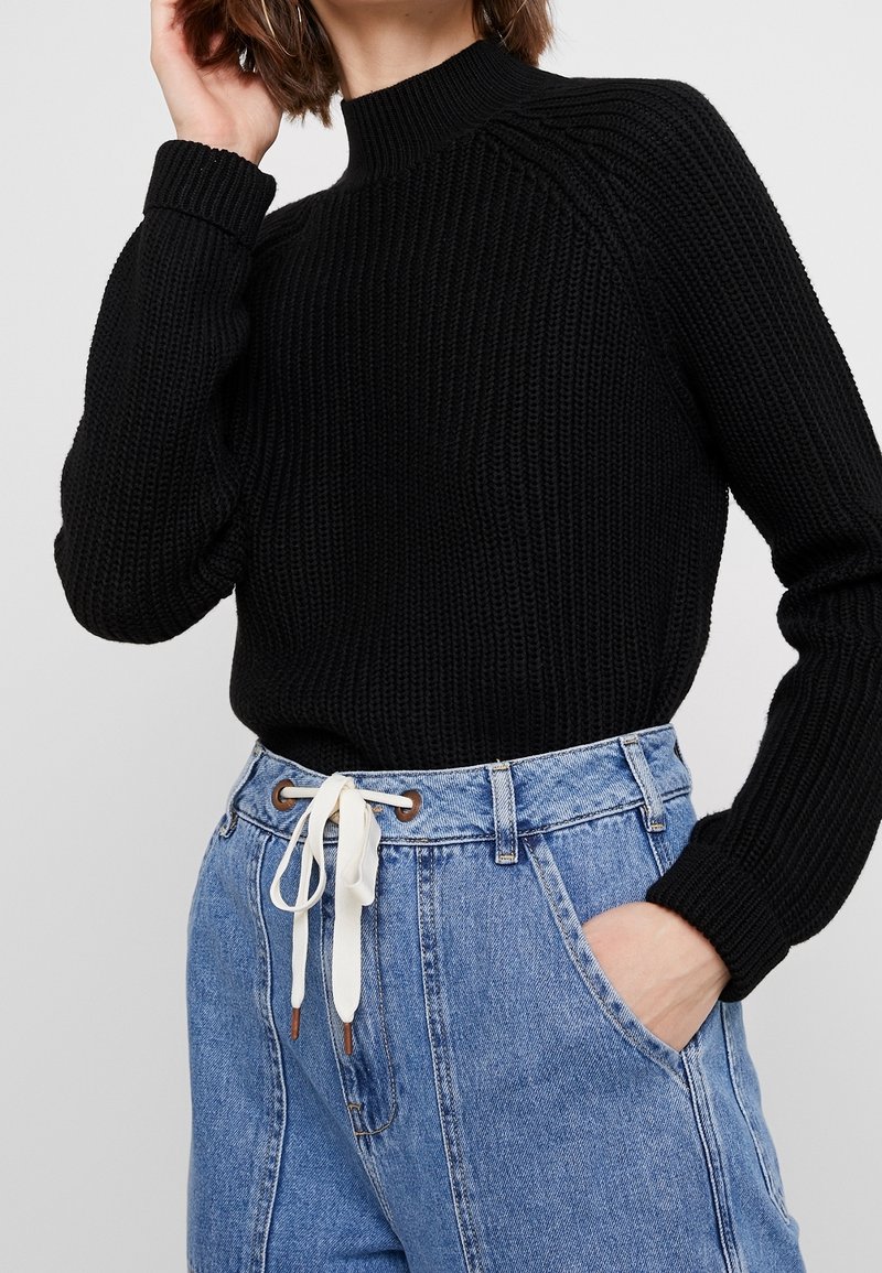 Black ribbed sweater with a high neck and long sleeves, paired with denim high-waisted trousers featuring a white drawstring and pockets.