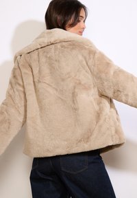 Beige nepbontjacket met een brede kraag, losse pasvorm en textuur. Zichtbare stiksels en een iets oversized model.