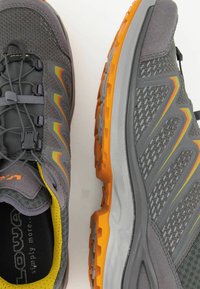 Graue Sport Schuhe mit einem strukturierten Mesh-Obermaterial, reflektierenden Akzenten in Gelb und Orange sowie einer robusten orangen Laufsohle.