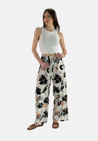 Witte crop top gecombineerd met loszittende, gebloemde broeken met grote zwarte bloemen en oranje accenten. Bruine sandalen maken de look af.