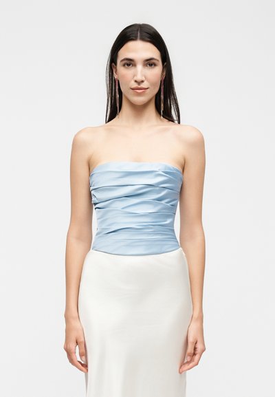 Femme aux longs cheveux foncés portant des boucles d'oreilles pendantes, un top bleu clair sans bretelles froncé et une jupe blanche, debout devant un fond clair uni.