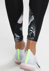 Piernas inferiores vestidas con leggings negros con paneles laterales abstractos multicolores y zapatillas deportivas blancas con lengüetas en el talón de un verde neón.