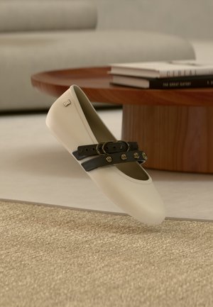 Chaussure plate beige avec deux sangles noires cloutées et boucles rondes, reposant sur un tapis beige, avec une table basse en bois et des livres en arrière-plan.