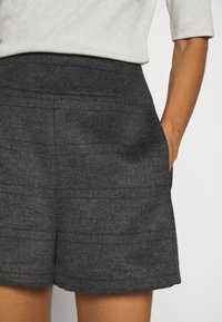 Femme portant un short en laine gris foncé avec des poches latérales et un haut de couleur claire, une main placée dans une poche.