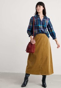 Zirņu raksta krekls tumši zilā, tirkīza un bordo krāsā; sinepju krāsas korduroja garā svārki; sarkana zamšādas soma; melnas pieguļošas potīšu zābaki.