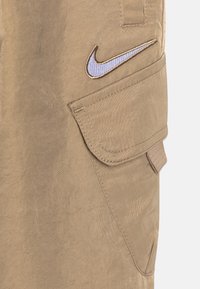 Béžové cargo kalhoty s texturovanou látkou, bočními kapsami a modrým vyšitým logem Nike na horní levé straně.