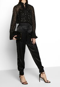 Svart genomskinlig blus med glittrande leopardfläckar, rynkad halsringning; kombinerad med svarta satinbyxor med sidoknappar och uppvikta fållar.