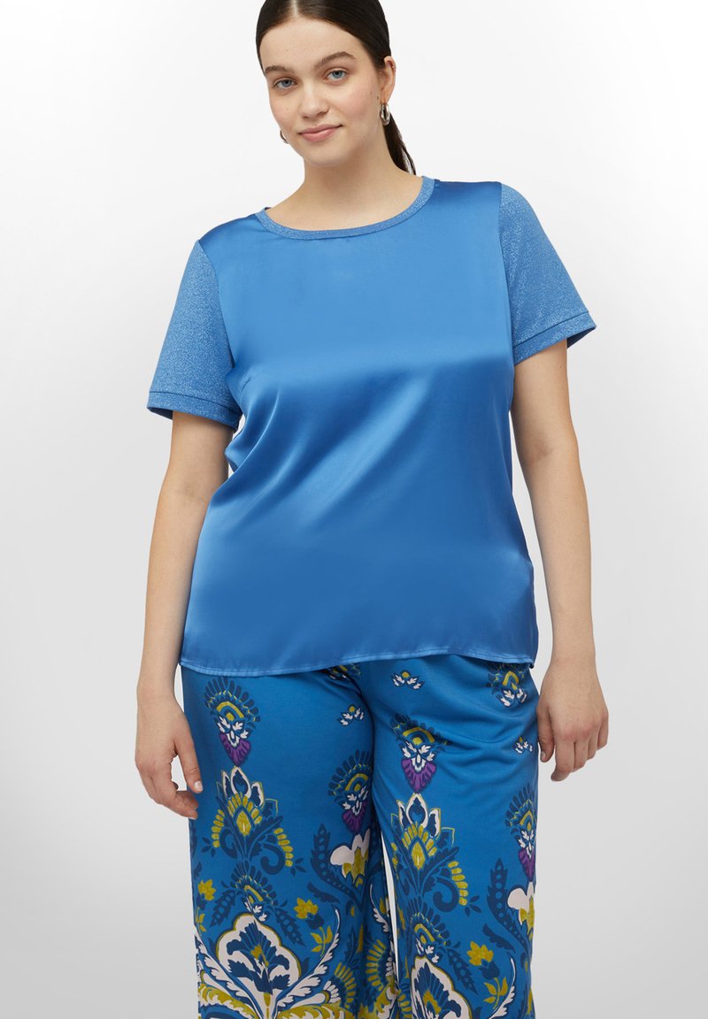 Top din satin albastru cu decolteu rotund și mâneci scurte, asociat cu pantaloni vibranți albaștri, cu un model floral în diverse culori.