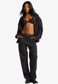 Sorte bukser med en løs pasform, parret med en sort bikini-top og en mørk knap-opp shirt. Modellen poser med hænderne i lommerne.