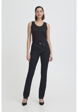 Žena stoji na jednostavnoj pozadini noseći crni top bez rukava, tamnoplave traperice s visokim strukom i crne štikle s šiljastim vrhovima.