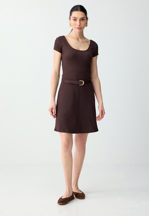 Femme portant une robe marron ajustée à manches courtes avec un col arrondi, ceinture marron avec boucle dorée, ballerines marron et boucles d'oreilles créoles dorées, debout.