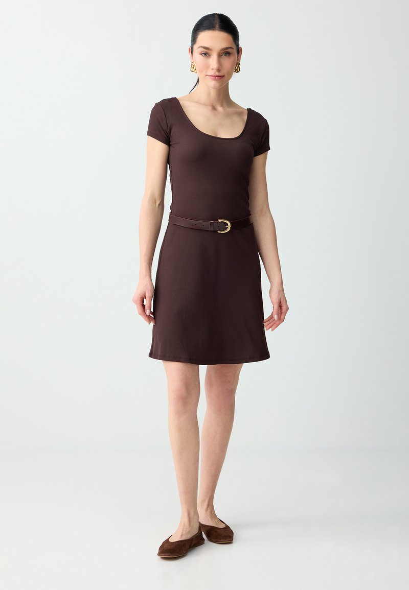 Femme portant une robe marron ajustée à manches courtes avec un col arrondi, ceinture marron avec boucle dorée, ballerines marron et boucles d'oreilles créoles dorées, debout.