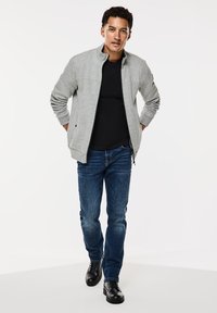 Light gray zip-up sweatshirt with ribbed cuffs, worn over a black shirt. Paired with blue jeans and black lace-up shoes. White background.