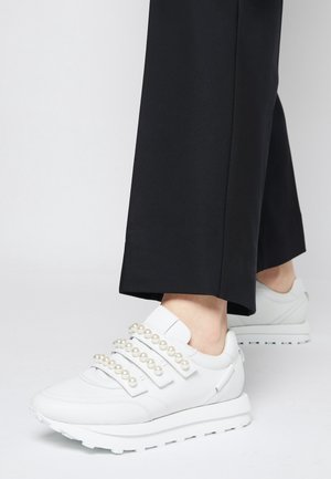 Hvide sneakers med perlebesatte stropper båret med sorte korte bukser, vist på en ensfarvet lys baggrund.