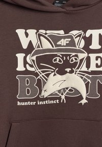 Chat portant un chapeau seau tenant un poisson dans la bouche, texte "instinct de chasseur" en dessous sur fond en tissu marron.