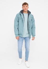 Lys blå vandtæt jakke med grå fleece-foring, frontknapper og justerbar hætte, båret over en lys blå skjorte og jeans.