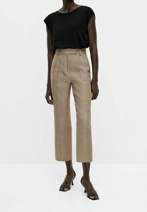 Woman wearing high-waisted beige cropped trousers, black sleeveless top, and black open-toe heeled sandals standing against plain background.