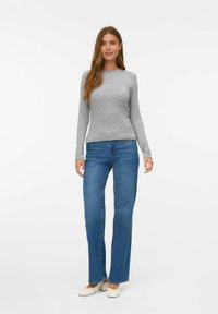 Vrouw staat in een grijze lange mouwen shirt, blauwe rechtlopende jeans en witte lage hak schoenen voor een witte achtergrond.