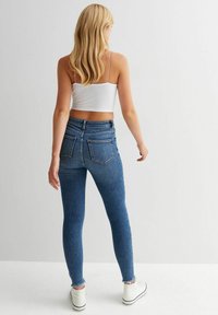 Kvinna med långt blont hår, klädd i en vit crop top, högmidjade blå tajta jeans och vita sneakers, står vänd bort.