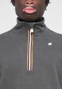 Fleece gris zippé avec col montant, présentant un détail de rayures multicolores et un petit logo sur la poitrine. Texture douce, design décontracté.