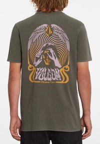 T-shirt vert olive avec un design graphique représentant un crâne, des mains et des ailes en violet et or, centré au dos. Manches courtes.