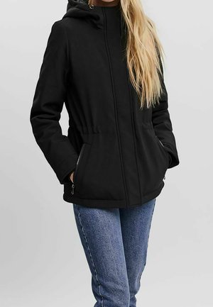 Femme portant une veste noire à capuche avec des poches zippées et un jean bleu, debout les mains dans les poches devant un fond uni.