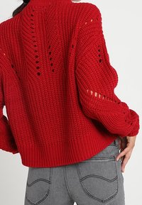 Pull rouge en maille avec un motif texturé, présentant des détails en maille ajourée sur les manches et un ourlet côtelé. Porté avec un jean gris.