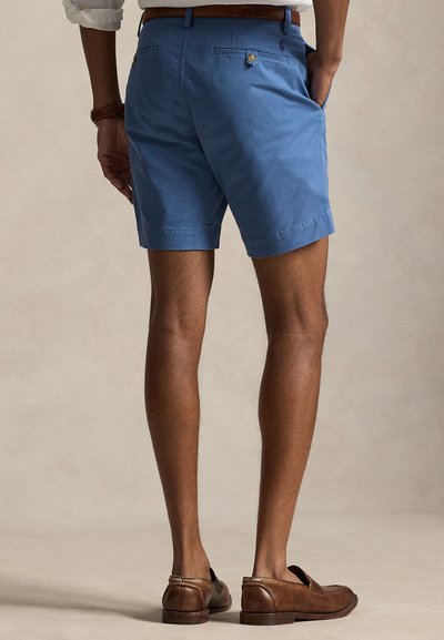 Man draagt blauwe op maat gemaakte korte broek, bruine leren riem, wit overhemd met opgerolde mouwen en bruine leren loafers, staand met de rug naar de camera.