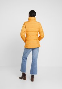 Kvinna som står vänd bort, iklädd en ljus orange puffjacka, ljusblå cropped jeans och mörkbruna klackstövlar.