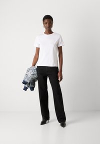 Femme debout sur fond blanc, portant un t-shirt blanc, un pantalon noir, des chaussures noires pointues, tenant une veste bleue à fleurs dans la main gauche.