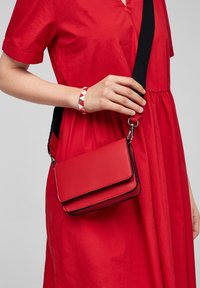 Rød crossbody taske lavet af tekstureret materiale med flaplukning, kontrasterende sort kant og justerbar rem.