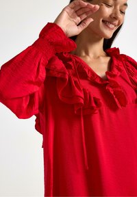 Rød bluse med flæser ved halsen og ærmerne, med snoretræk detalje og et tekstureret stof, der viser en afslappet pasform.