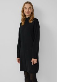 Schwarzes Strickkleid mit hohem Kragen, überschnittenen Schultern und langen Ärmeln. Locker geschnitten mit gerippten Bündchen. Minimalistisches Design.