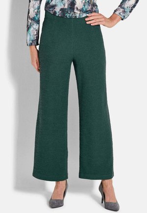 Wijdlopende broeken van donkergroene stof met een gestructureerd oppervlak; gecombineerd met een gebloemde blouse en grijze hakken.