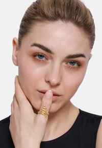 Goldener ring in Schlangenform mit strukturierten Schuppen, an einem Finger getragen; das Model hat einen neutralen Gesichtsausdruck und hält die Hand nah am Gesicht. Schwarzer Hintergrund.