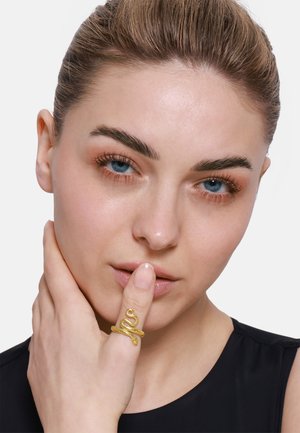 Goldener ring in Schlangenform mit strukturierten Schuppen, an einem Finger getragen; das Model hat einen neutralen Gesichtsausdruck und hält die Hand nah am Gesicht. Schwarzer Hintergrund.
