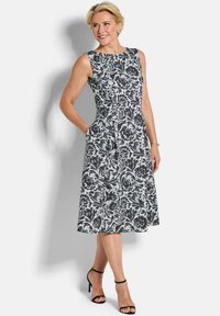 Vrouw in een mouwloze zwart-witte midi-jurk met bloemenprint en zijzakken, met zwarte enkellaarzen met enkelbandje, staande tegen een witte achtergrond.