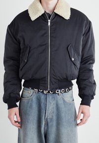 Chaqueta bomber negra con cuello de pelo sintético beige en contraste, cierre de cremallera en el frente y bolsillos laterales. Combinada con jeans de mezclilla azul de tiro alto.