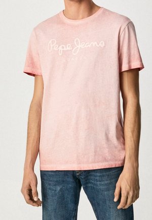 Muž v světle růžovém tričku s krátkým rukávem s nápisem "Pepe Jeans London" a modrých džínech na jednobarevném pozadí.