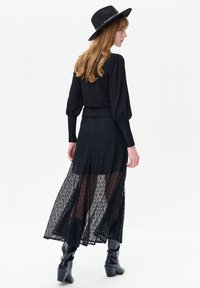 Zwarte blouse met lange mouwen en opgestikte mouwen, gecombineerd met een zwarte maxirok met laagjes kant. Model draagt ook een zwarte brede hoed en kniehoge laarzen.