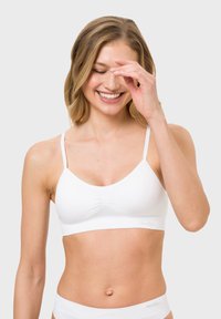 Weißes Bralette mit dünnen, verstellbaren Trägern, nahtlosem Design, weichem Stoff und gerafftem Detail vorne. Kombiniert mit weißer Unterwäsche.