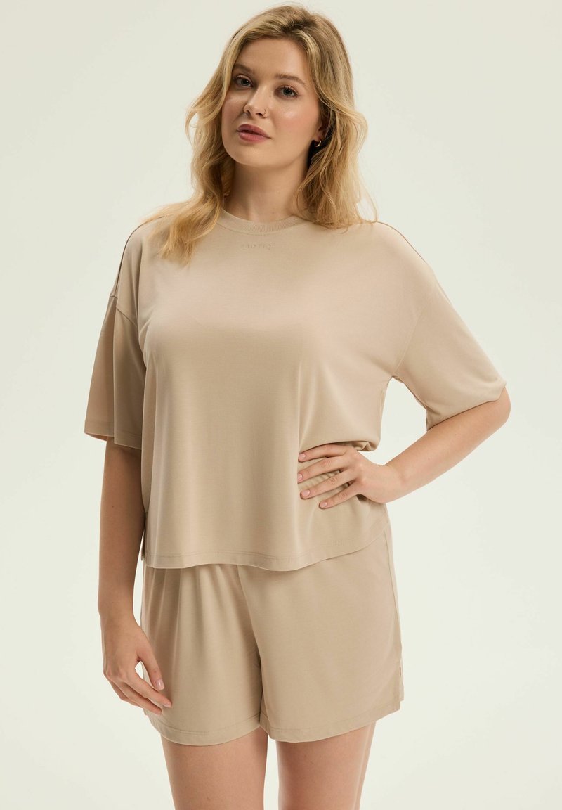 Beżowa bluzka na krótki rękaw o luźnym kroju, z okrągłym dekoltem i subtelnym logo; współtworzy z dopasowanymi szortami, wykonanymi z miękkiego materiału.