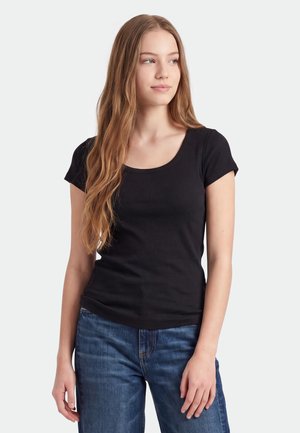 Junge Frau mit langen hellbraunen Haaren, die ein schwarzes kurzärmeliges Oberteil und blaue Jeans trägt, schaut seitlich vor einem schlichten Hintergrund.