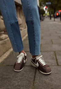 Rudi ir balti Nike sportbačiai su tekstūruotu viršumi, baltais raišteliais ir retro siluetu. Suderinti su šviesiai mėlynomis džinsomis su suplėšytu apvadu.
