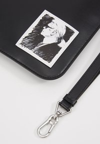 Sac en cuir noir avec bandoulière amovible et clip argenté, arborant une pièce blanche cousue avec un croquis en noir et blanc en profil d'une personne aux cheveux tirés en arrière.