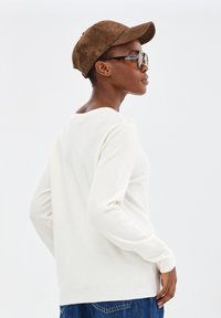 Vrouw in een witte lange mouwen shirt, blauwe jeans, bruine suede pet en schildpadprint zonnebril, die naar links kijkt op een effen witte achtergrond.