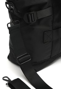 Crna najlon torba s podesivom naramenicom, metalnim dijelovima i detaljima od šivanja. Glatka tekstura s minimalističkim dizajnom.