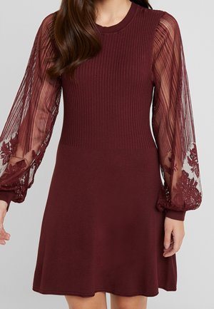 Woman wearing a burgundy dress with ribbed bodice, sheer long sleeves featuring floral lace embroidery, and fitted cuffs.
